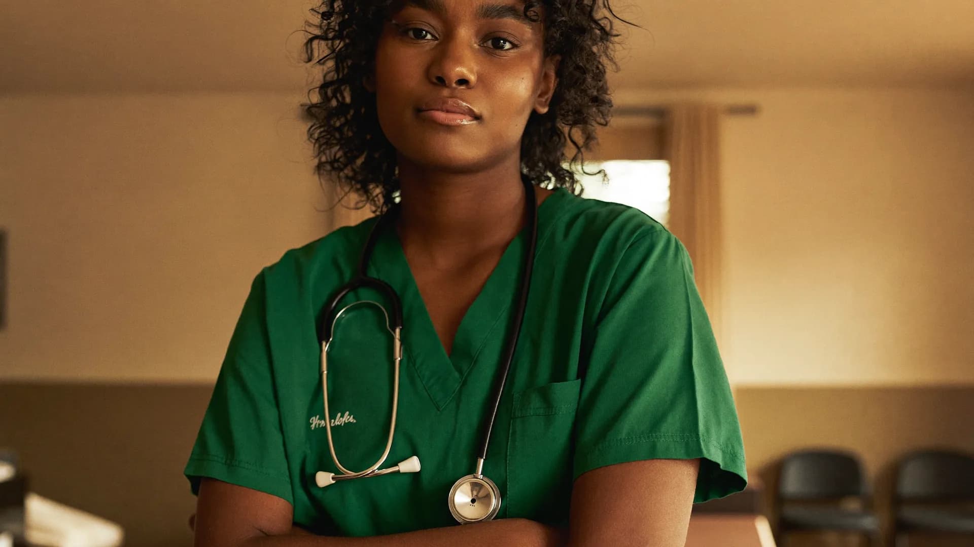 A young nurse in scrubs with a stethoscope around her neck, lifting her head with quiet confidence in a warmly lit training room