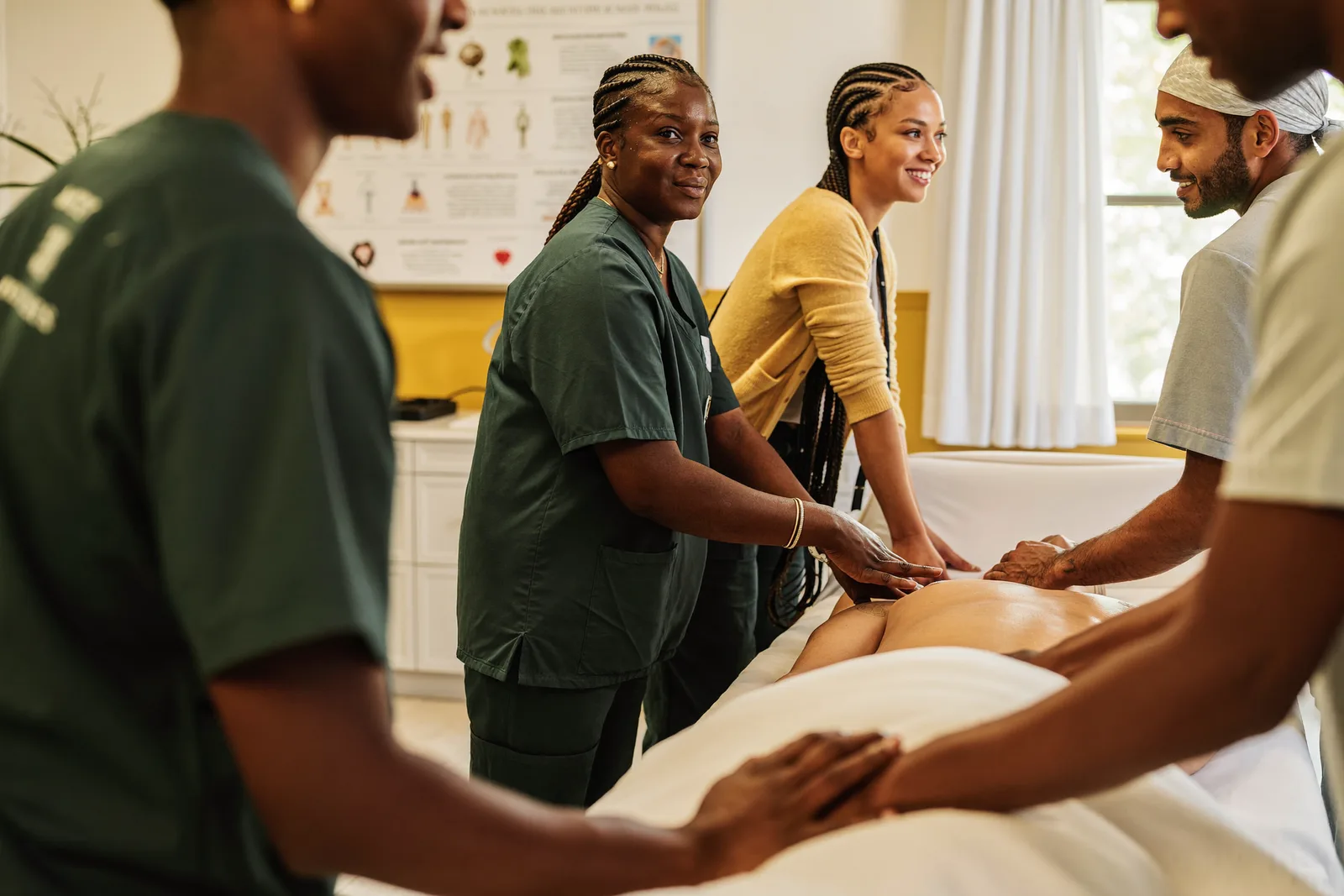 CNA students in scrubs practicing patient care skills on a training bed while an instructor demonstrates positioning technique